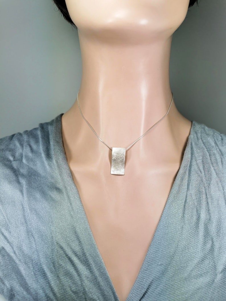 Puede incluir: Un collar de plata con un colgante rectangular. El colgante tiene una superficie texturizada.