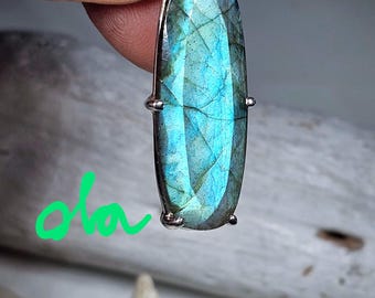May include: A silver pendant with a large oval-shaped labradorite gemstone. The gemstone is a deep blue-green color with iridescent flashes of light.