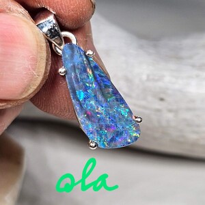 May include: A pendant featuring a vibrant, iridescent blue opal gemstone. The opal is teardrop-shaped, with flashes of green, purple, and pink. It is set in a silver-colored metal setting with small decorative accents. The word "ola" is written in green cursive.