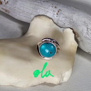 May include: A silver ring with a turquoise stone set in a curved band. The ring is on a white background with the word "ola" written in green.