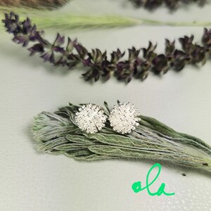 May include: A pair of silver stud earrings with a white, round, textured design. The earrings are on a green leafy background.