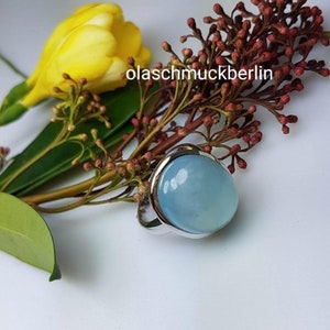 May include: A silver ring with a large, oval-shaped, light blue gemstone. The ring is surrounded by green leaves and red flower buds.