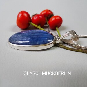 May include: A silver pendant with a blue gemstone set in an oval shape. The pendant is hanging from a silver chain. The pendant is surrounded by red berries.