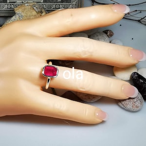 May include: A gold ring with a square, red gemstone. The ring is on a model's finger.