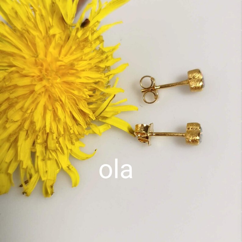 May include: A pair of gold stud earrings with small, round, textured gemstones. The earrings are shown against a white background with a yellow dandelion flower.