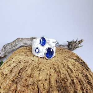 May include: A wide silver ring with a textured surface, featuring several blue gemstones of varying sizes. The gemstones are oval and round, set in a modern, artistic design. The ring is displayed on a piece of wood.