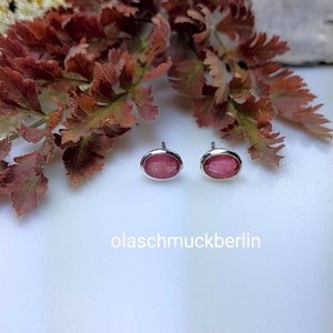 Op de afbeelding: Een paar ovale oorknopjes met roze edelstenen en zilveren zettingen. De oorbellen worden gepresenteerd op een wit oppervlak, met roodbruin blad op de achtergrond. De tekst "olaschmuckberlin" is onderaan zichtbaar.
