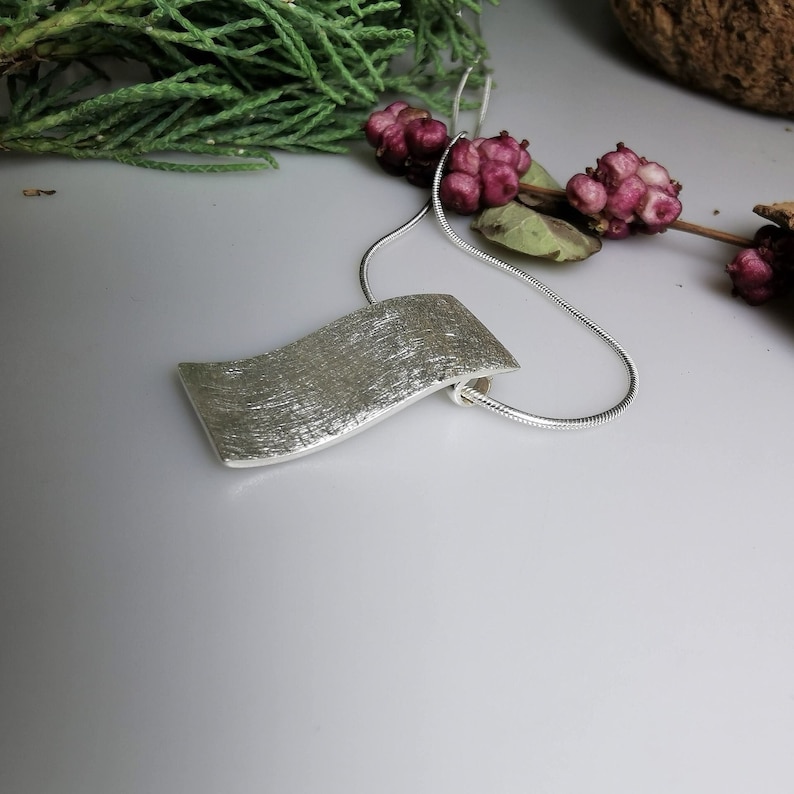 Puede incluir: Un collar de plata con un colgante ondulado texturizado. El colgante es un dise&ntilde;o simple y moderno.