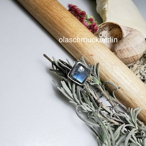 May include: A square-faced silver ring with a blue and green iridescent stone. The ring is set against a backdrop of dried herbs, a wooden dowel, and a white surface. The text "olaschmuckberlin" is visible.