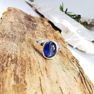 May include: A silver ring with a large, oval-shaped, deep blue gemstone. The ring is set on a piece of textured wood, showcasing the gemstone's color and the ring's design. The gemstone has a faceted surface.