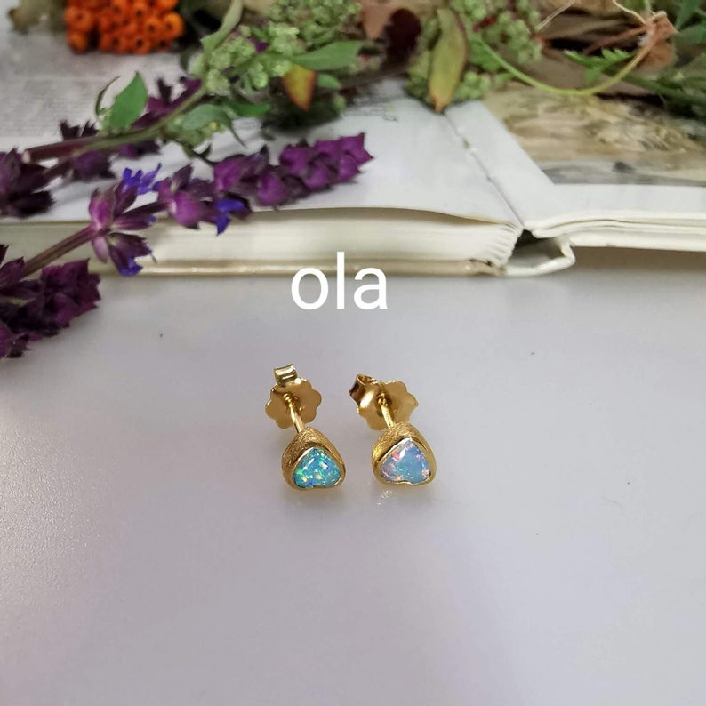 May include: A pair of gold stud earrings with opal stones. The earrings are set in a triangular shape. The opal on the left earring is a light blue color, while the opal on the right earring is a white color. The word "ola" is written in black text on a white background.