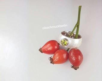Op de afbeelding: Een zilveren ring met een getextureerd oppervlak en een ronde, lichtgroene edelsteen. De ring ligt naast drie rode rozenbottels en een groene stengel. De tekst "olaschmuckberlin" is zichtbaar.