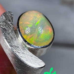 Könnte beinhalten: Nahaufnahme eines Silberrings mit einem großen, ovalen Opal-Edelstein. Der Opal zeigt ein lebendiges Farbspiel mit grünen, gelben und roten Farbtönen. Der Ring hat ein gebürstetes Metall-Finish.