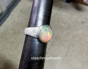 May include: A silver ring featuring a large, oval opal gemstone. The opal showcases a blend of colours, including pink, green, and yellow. The ring is set on a dark metal object. The text "olaschmuckberlin" is visible.