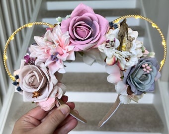 May include: A pair of light-up Minnie Mouse ears with a floral design. The ears are decorated with pink, white, and gray flowers, and feature a small figurine of a man and a child.