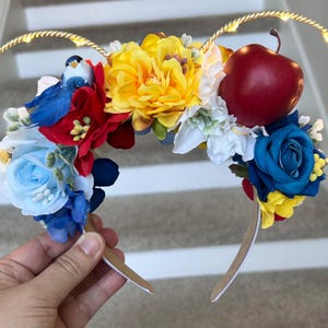 May include: A decorative headband with a red apple, blue and yellow roses, and a small blue bird. The headband has gold-coloured hoops with small lights. The headband is held by a hand.