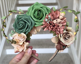 May include: A pair of light-up floral headband ears with a small green figurine in the center. The headband is decorated with green, pink, and brown flowers, and has a copper wire vine with lights wrapped around it.