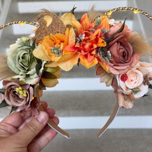 May include: A headband featuring two circular ears outlined in twisted gold metallic cord with integrated warm white LED lights. The headband is decorated with an arrangement of artificial flowers in shades of sage green, orange, and dusty rose.