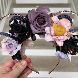 May include: A decorative headband featuring a floral arrangement with black, purple, and pink roses. The headband has a twisted wire frame with small lights and is accented with decorative elements.