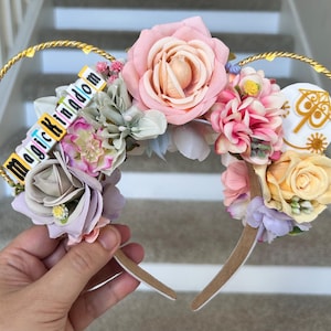 Può includere: Un paio di orecchie di topo bianche con un cerchietto dorato decorato con fiori rosa, bianchi e gialli. Il cerchietto presenta anche un ciondolo bianco con un design dorato e il testo "Magic Kingdom" in lettere colorate.