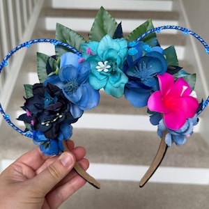 May include: A headband featuring two circular wire frames wrapped in blue metallic cord, adorned with an arrangement of artificial flowers in shades of navy, royal blue, turquoise, and a single vibrant hot pink blossom, accented by green leaves.