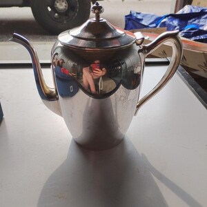 May include: A polished silver-toned metal teapot featuring a curved spout, a sturdy handle, and a removable lid with a small finial knob on top. The reflective surface of the teapot displays a distorted view of the surrounding environment.