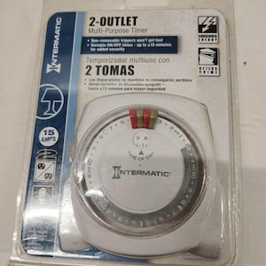 Op de afbeelding: Een Intermatic 2-stopcontact tijdschakelaar in de originele plastic verpakking. Het witte ronde apparaat heeft een 24-uurs wijzerplaat met rode en groene schakelruiters, is geschikt voor 15 ampère en bevat tekst over variabele in- en uitschakeltijden.