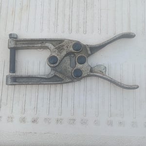 May include: A silver-toned metal hand tool featuring a lever-action mechanism with four dark circular rivets. The tool has two curved handles and a threaded bolt with a nut at the end, positioned against a white surface with imperial markings.