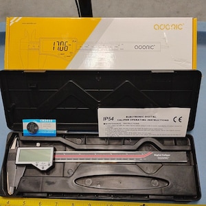 Peut inclure: Un pied à coulisse numérique Adoric dans un étui de rangement en plastique noir avec une boîte jaune, une pile au lithium CR2032 et un manuel d'instructions. Un mètre ruban jaune est placé sous l'étui, indiquant une longueur de 38 centimètres.