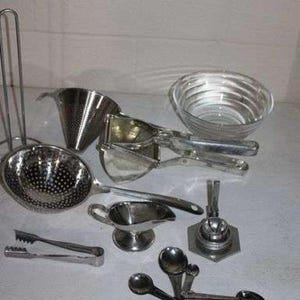 May include: A collection of stainless steel kitchen utensils including a conical strainer, a potato ricer, a small gravy boat, a set of measuring spoons, a pair of tongs, a small glass bowl, and a metal stand, all arranged on a flat surface.