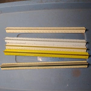 May include: A collection of five triangular architectural scale rulers in various colors including off-white, white, and yellow. Each ruler features multiple graduated measurement markings along its three sides, designed for technical drafting and engineering.
