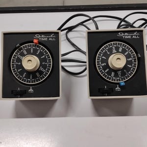 Puede incluir: Dos temporizadores eléctricos Intermatic Time All con carcasas cuadradas de color beige y paneles de control negros. Cada unidad cuenta con un dial circular de 24 horas con marcas AM y PM, una perilla central beige y un interruptor deslizante.