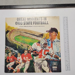 Può includere: Una copertina di un disco in vinile intitolata Great Moments in Ohio State Football, un tributo a Woody Hayes. La copertina presenta un'illustrazione dipinta di uno stadio di football e giocatori, con un righello che mostra un'altezza di circa 31 cm.