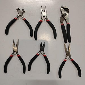 May include: A set of six metal pliers with black rubberized handles and red accents, including end cutters, needle-nose, and slip-joint pliers, arranged above a ruler showing measurements in inches along the bottom edge of the frame.