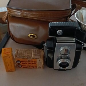 May include: A black and silver Kodak Brownie Reflex 20 camera with a flash attachment, a brown leather carrying case, a yellow Kodak VP 620 film box, and two packages of General Electric M2 flash bulbs with printed exposure charts on the front.