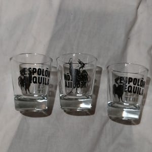 May include: Three clear glass shot glasses with thick bases. Each glass features a black graphic print of a rooster and the text ESPOLON TEQUILA. The glasses are arranged in a row, displaying the brand logo and the bird illustration clearly.