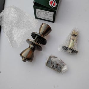 Pode incluir: Um conjunto de fechadura de privacidade em latão antigo com dois puxadores redondos, um mecanismo de trinco e uma chapa testa. A caixa está rotulada como Privacy Lock, US5 Antique Brass, RIZ-20 e Cal-Royal Products, Inc. Componentes embalados.