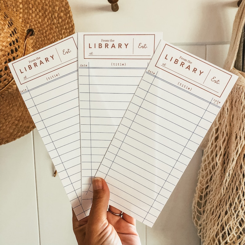 May include: Three white library cards with a vintage design are held in front of a neutral background. Each card has the text "From the LIBRARY Est." at the top, with spaces for "of" and "(title)" and a date. The cards are lined.