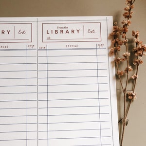 May include: A white library record sheet with blue lines and brown text. The text reads "From the LIBRARY" and "Est." The sheet has space for titles and ratings. A sprig of dried flowers is in the background.