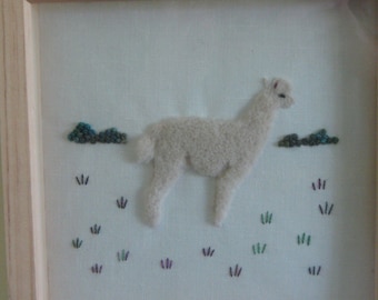 May include: A framed textile artwork depicting a white llama in a field. The llama is made of textured material, set against embroidered grass and small blue and green cloud-like shapes. The frame is made of light-coloured wood.