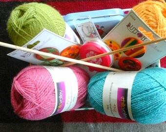 May include: A selection of knitting supplies, including balls of yarn in green, pink, and teal, along with orange and red buttons, a blue tin, and knitting needles. The yarn is labeled as 50g.