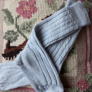May include: A pair of pale blue ribbed knit socks. The socks are folded on a floral cushion with pink and green detailing. The socks have a traditional design.