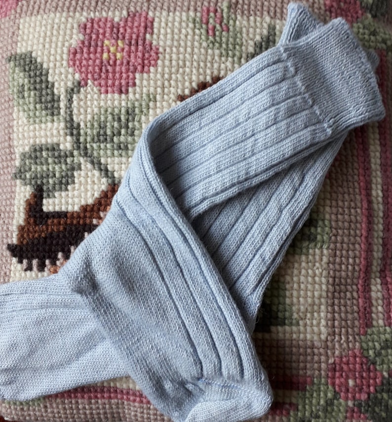 May include: A pair of pale blue ribbed knit socks. The socks are folded on a floral cushion with pink and green detailing. The socks have a traditional design.