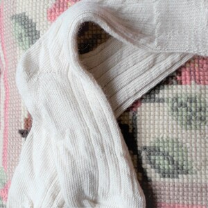 May include: A pair of cream-coloured ribbed knit socks. The socks are folded over, displaying the ribbed texture and the heel. The fabric looks soft and comfortable. The background is a patterned rug with pink, white, and grey elements.
