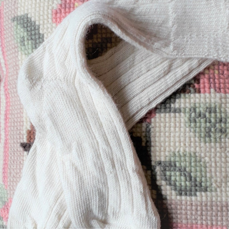 May include: A pair of cream-coloured ribbed knit socks. The socks are folded over, displaying the ribbed texture and the heel. The fabric looks soft and comfortable. The background is a patterned rug with pink, white, and grey elements.