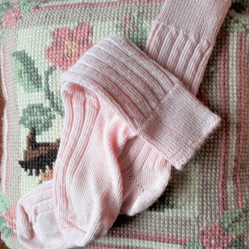 May include: A pair of pale pink, ribbed knit socks with a folded cuff. The socks are made of a soft, textured material. The socks are lying on a pink, green, and white patterned surface.