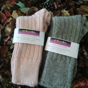 May include: Two pairs of ribbed knit socks, one in pale pink and the other in grey, are shown. Each pair is wrapped with a white label that reads "chillavalley Alpaca Bed Socks." The socks are made of 90% British Alpaca and 10% Nylon, and are a small size.