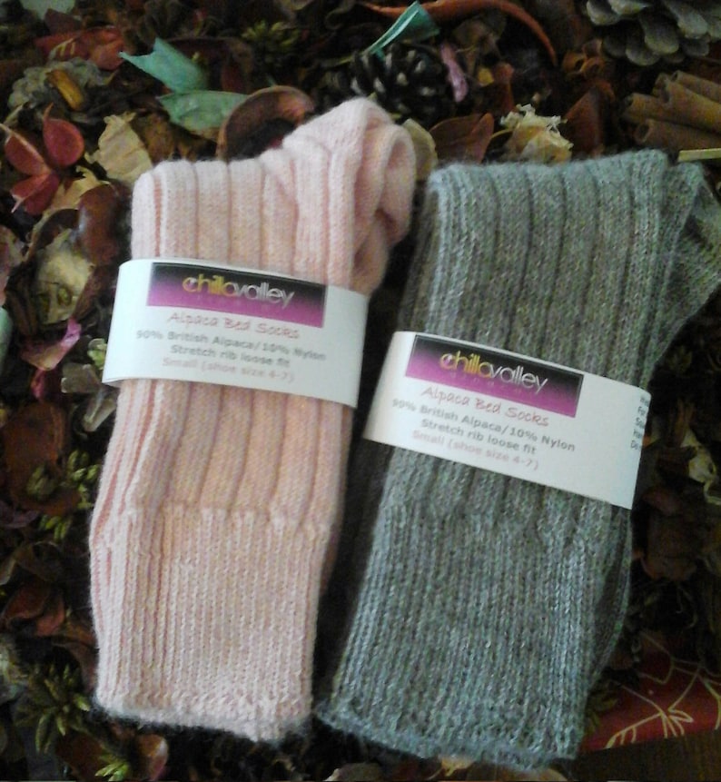 May include: Two pairs of ribbed knit socks, one in pale pink and the other in grey, are shown. Each pair is wrapped with a white label that reads "chillavalley Alpaca Bed Socks." The socks are made of 90% British Alpaca and 10% Nylon, and are a small size.