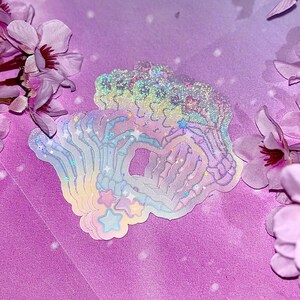 May include: A set of iridescent rainbow holographic stickers featuring a skeleton hand design with stars. The stickers are layered on top of each other and have a glittery finish.