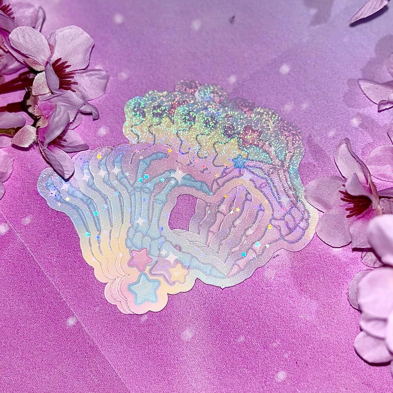 May include: A set of iridescent rainbow holographic stickers featuring a skeleton hand design with stars. The stickers are layered on top of each other and have a glittery finish.